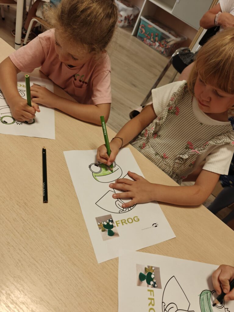 Dzieci przy stoliku kolorują prace plastyczną - żaby.